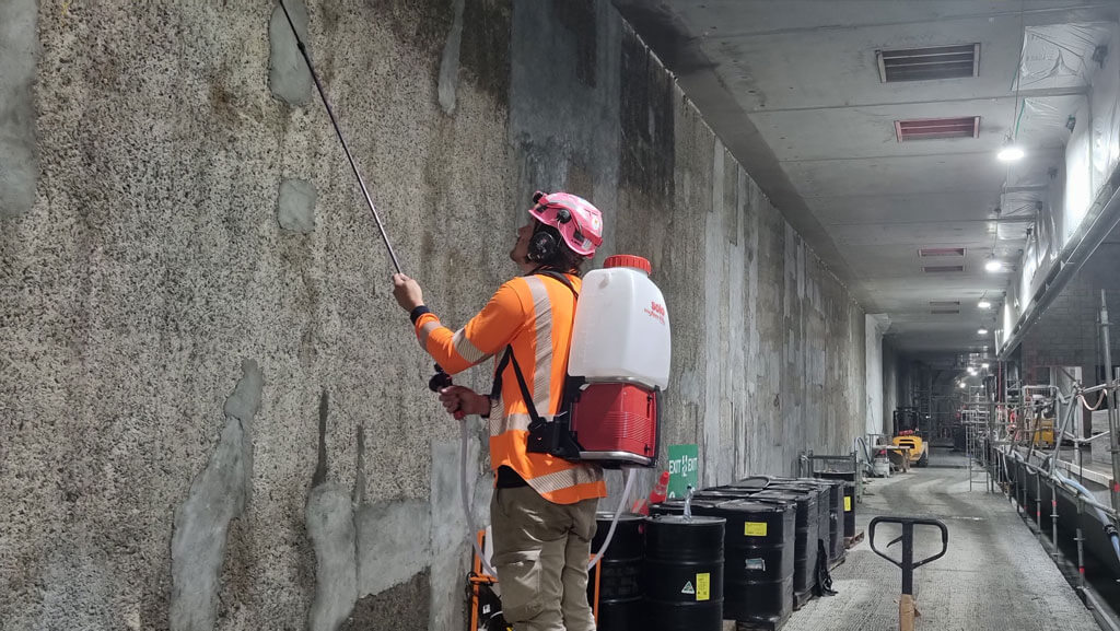 A worker in high-visibility clothing applies concrete waterproofing treatment to a wall in an industrial or construction setting using a sprayer backpack.